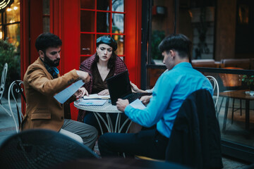 Three business professionals engage in a focused meeting at a quaint outdoor cafe, discussing documents and working on a laptop. The atmosphere is serious and collaborative, fostering creative