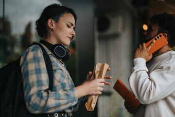 Two students socializing outdoors during a college break. One student is holding a snack while the other talks on a phone, capturing a moment of casual conversation and relaxation.
