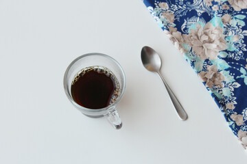 Top-down view of transparent glass coffee cup and silver spoon on white table with blue fabric napkin on top right corner. Hot soft drink, caffeine, stimulating beverage.