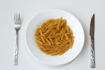 Crockery plate with uncooked penne pasta and cutlery on the table. Raw italian gluten food on white dish. Dinning, hunger, meal, diet concept.