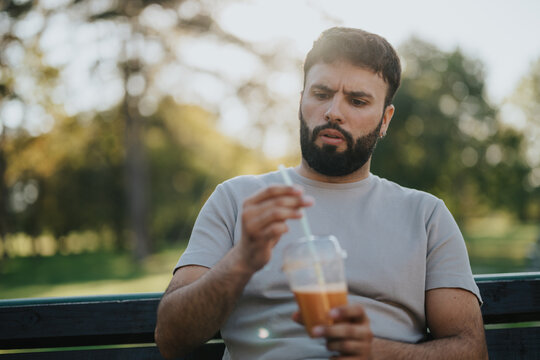 A thoughtful man sits on a park bench examining his drink, pondering its taste or ingredients. The serene outdoor setting enhances the contemplative mood, making this a candid capture of curiosity. - Powered by Adobe