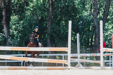 A young rider confidently guides a horse through an obstacle course in a lush, green outdoor setting. The equestrian wears safety gear and displays skillful control.