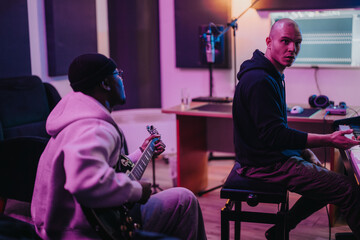 Two musicians working together in a recording studio, one playing guitar and the other on keyboard. The artistic setting features modern equipment and creative ambiance, promoting innovation and