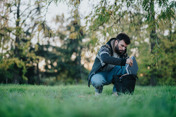 A man kneels on grass, packing a laptop into a backpack under lush green trees. The peaceful park environment suggests a productive and tranquil outdoor work setting.