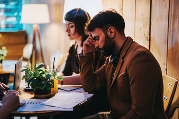 A tired business professional sits in a cafe, visibly stressed, working late with papers and a laptop. The cozy environment contrasts with his fatigue and concentration.