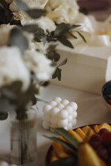 A white candle stands on a table next to a vase of flowers. Wedding accessories. Background for a wedding or event invitation.