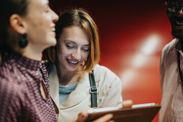 A diverse group of colleagues engaging in a collaborative discussion with a tablet in a creative office setting, showcasing teamwork and enthusiasm against a bold red backdrop.