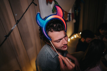 A young man enjoys a lively birthday party with friends, wearing colorful devil horns in a festive setting. The celebration is filled with laughter and joy.