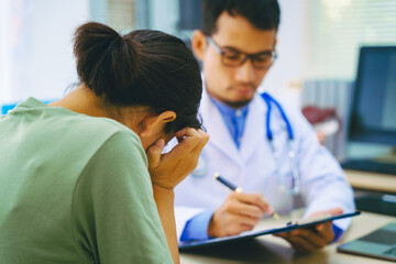 A female patient meets a male doctor at a desk in a hospital, discussing her migraine headache,...