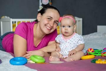 Obraz premium A mother and her adorable baby girl are enjoying playtime on a cozy bed. Surrounded by colorful toys, they share smiles and laughter in a warm, inviting atmosphere.