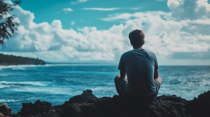 Fototapeta premium A man sitting on a rock by the ocean, looking out at the horizon
