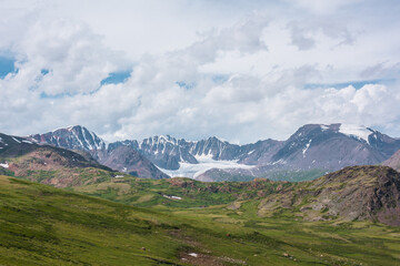 Scenic alpine landscape with green hills and rocks with view to big glacier and large snow-capped mountain range far away under clouds in blue sky in changeable weather. Awesome high snowy mountains.