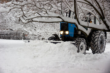 A blue tractor clears snow in a city park after heavy snowfall. taken during cleaning, in winter, during the day, in sunny weather
