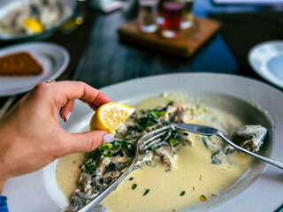 Golden trout in cream sauce in white plate at restaurant. Hand holding a slice of lemon on a plate with fish and tableware.