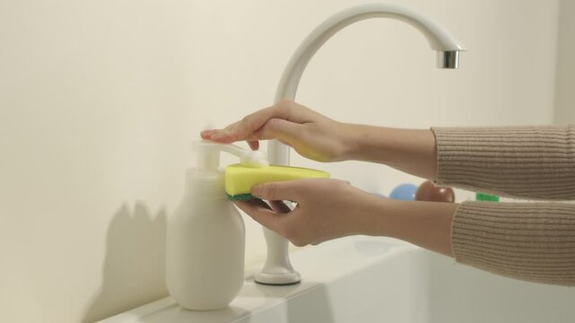Template video about cleaning product, frontal view of a model pushing liquid from a white container mockup into a sponge, placed next to some toys, in the kitchen setting.