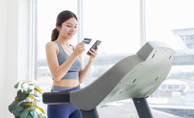 Asian young beautiful woman, sporty active energetic girl in sportswear exercise indoors using smartphone and credit card jogging on treadmill training in living room, health care wellness lifestyle.