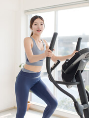 Fototapeta premium Asian young beautiful cheerful happy woman active girl in sportswear stationary exercise bike movement speed energy training in fitness gym or living room at home, healthy wellness action lifestyle
