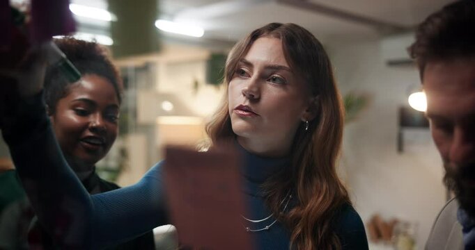 Business people, meeting and writing with sticky note at night for schedule planning, project or brainstorming at office. Group, employees or team with reminder for agenda on glass board at workplace