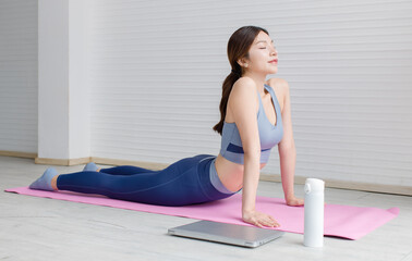 Asian young beautiful attractive woman wearing sportswear stretches body upward facing on mat, yoga workout pilates exercise training with laptop and water bottles in living room healthy wellness.