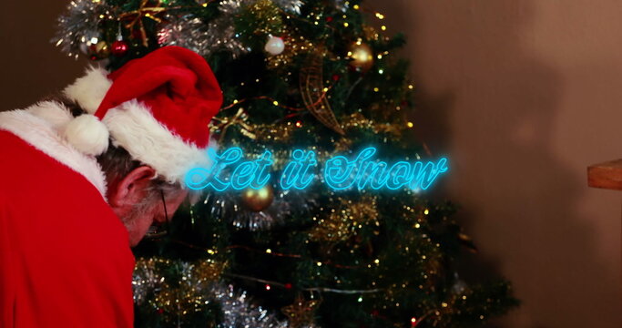 Image of neon blue let it snow text banner over santa claus decorating a christmas tree