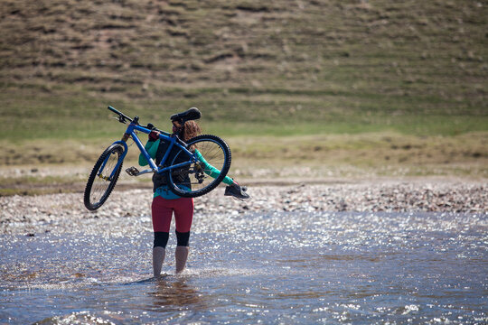 Woman carrying bike through shallow water on a sunny day.