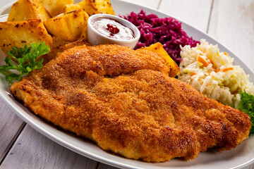 Crispy breaded fried pork chop, fried potatoes, ketchup and red and white cabbage served on white plate on wooden table