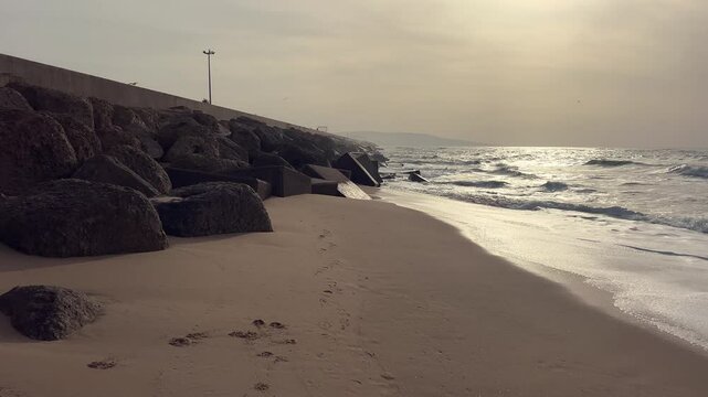 Gentle waves washing onto the sandy shore at sunset, with footprints leading along the beach and a breakwater in the background in barbate, andalusia, spain