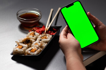 Female using smartphone while eating sushi rolls at home. Smartphone with green screen with space for text. Smartphone in hands with served table in background.