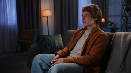 Young man spending evening at home sitting on sofa in living room, switching tv with remote watching horror movie, scared shocked expression.