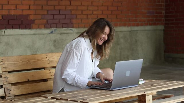 Smiling blond businesswoman in white blazer sitting on wooden pallets  using typing on laptop computer and have conversation online gesticulating, studying outdoors. Female psychologist working remote