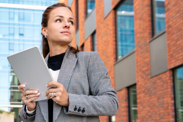 Businesswoman with digital tablet stands outdoors in an urban district next to a business buildings.