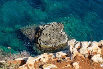 a dangerous view from above the cliffs into the clear water of the coastline of Comino, Malta. With an big rock in the middle.