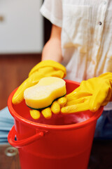 Woman in yellow rubber gloves holds a sponge in a red bucket with a yellow sponge on top on a wooden table