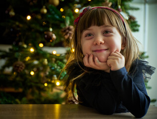 Smiling child leans on a table by a decorated Christmas tree full of expectation and anticipation....