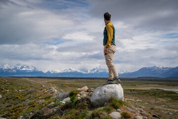 A man stands atop a rock, gazing at the vast Patagonian wilderness with snow-capped mountains in...