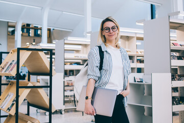 Half length portrait of successful Caucasian female freelancer with laptop computer posing in public bookstore, youthful student 20 years old looking at camera near bookshelf in coworking space