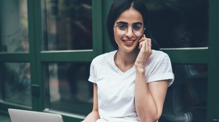 Portrait of successful female freelancer enjoying cellphone conversation during remote work at urban setting, happy Hispanic woman with laptop computer making international phone call for communicate