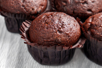Fresh chocolate muffins with oily texture and deep cracks in brown paper cups on a light wooden aged background.