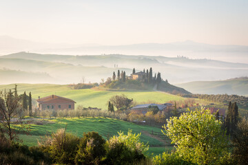 Rolling hills of Tuscany at sunrise, bathed in soft mist in Spring. A serene farmhouse surrounded by cypress trees and lush greenery creates a dreamy, picturesque Italian countryside landscape