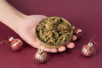 in a woman's hand a bowl with dry buds of medical marijuana, surrounded by Christmas and New Year's decorations balls on a dark red background