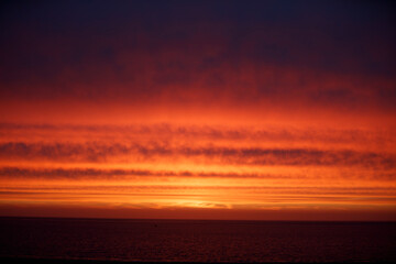 Vibrant sunset over a calm ocean; fiery orange and dark clouds.  Ocean's dark hues mirror the sky's...