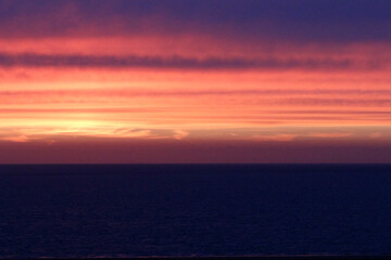 A vibrant sunset over a dark ocean.  Warm colors transition to cool as day fades.  Calm seas reflect the fiery sky.