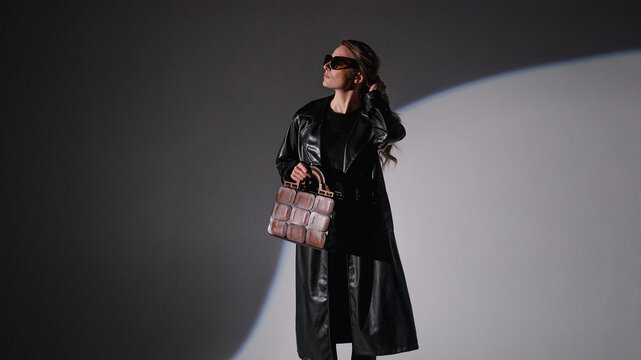 A fashionable woman in a black leather coat and sunglasses poses against a gray background with a spotlight. She carries a unique brown handbag and looks off to the side. 