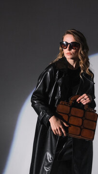 A woman with blonde wavy hair, wearing sunglasses and a black leather trench coat, holds a unique, structured brown handbag. She poses in a studio setting with a spotlight effect. 