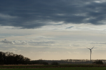 Silhouetted wind turbine against a cloudy, overcast sky.  Trees line the horizon.  A tranquil rural scene.