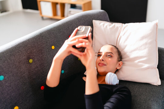 A young ballerina with white headphones rests on a gray couch, holding her phone above her, with a pink curtain and modern vibrant decor creating a bright indoor atmosphere.