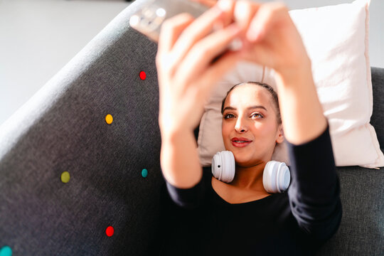 A young ballerina with white headphones rests on a gray couch, holding her phone above her, with a pink curtain and modern vibrant decor creating a bright indoor atmosphere.