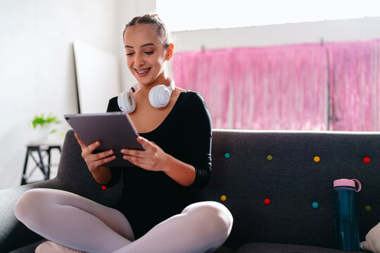 A young ballerina sits cross-legged on a gray couch, wearing white headphones and using a tablet, with a pink curtain and vibrant modern decor visible in the background.