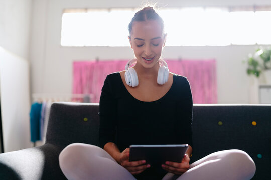 A young ballerina sits cross-legged on a gray couch, wearing white headphones and using a tablet, with a pink curtain and vibrant modern decor visible in the background.