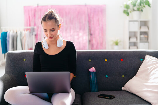 A young ballerina with headphones around her neck uses a laptop while sitting on a gray couch, with a bright studio environment featuring pink curtains and greenery in the background.